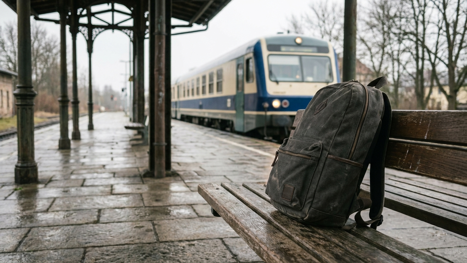 Eine schlichte Laptoptasche aus dunklem Stoff steht auf einer Bahnsteigbank, im Hintergrund unscharf ein einfahrender Regionalzug