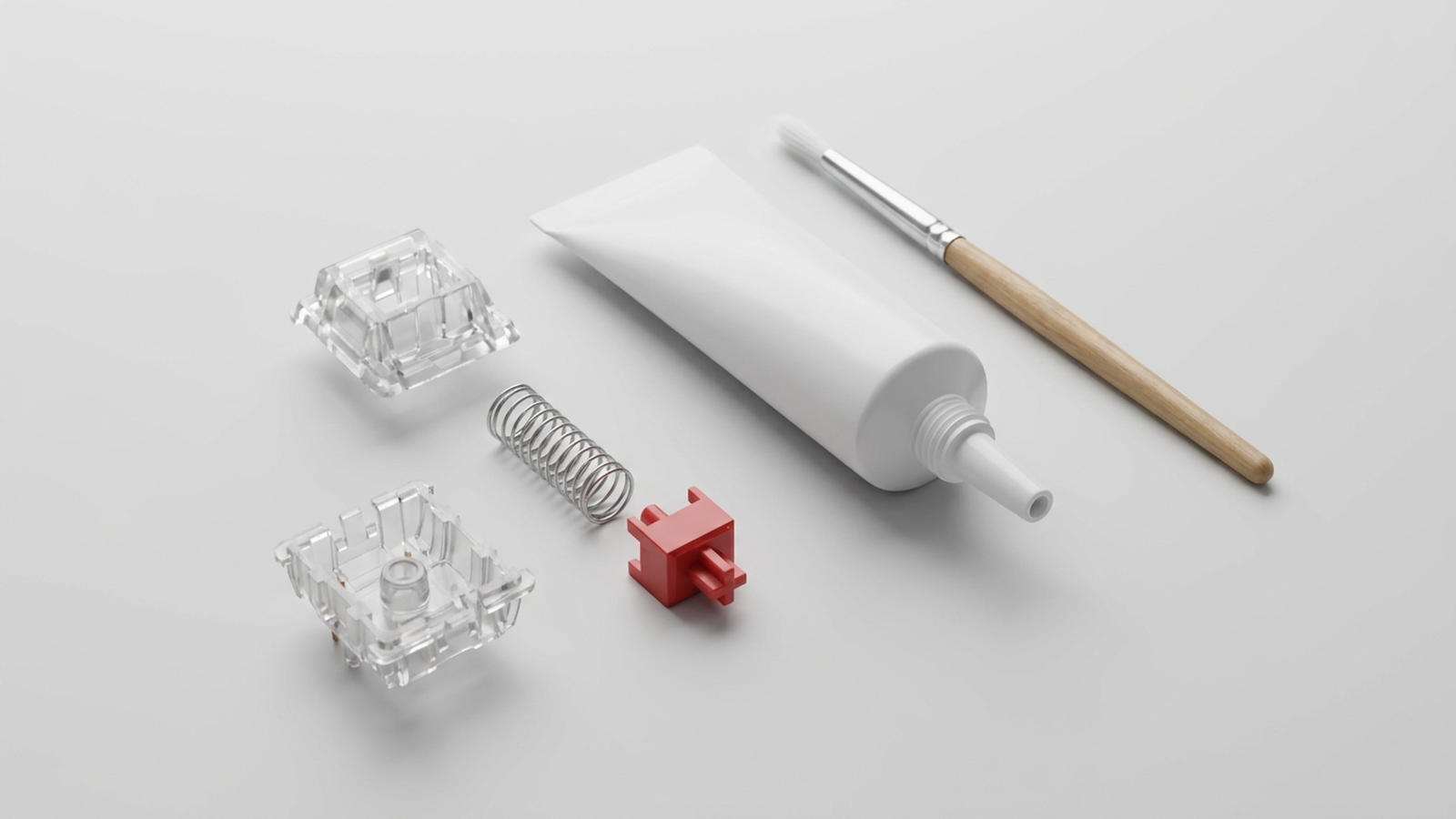 Disassembled mechanical keyboard switch housings, a spring, a stem, a tube of switch lubricant, and a small brush laid out on a white surface